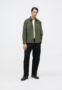 Camisa de botones verde oliva con dos bolsillos frontales, llevada sobre una camisa blanca, combinada con pantalones negros y zapatos oscuros.