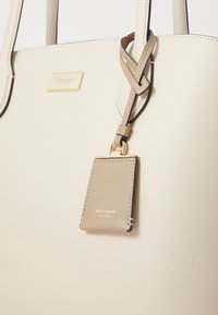 Beige tote bag with textured leather finish, featuring a gold logo plaque and a matching tag attached by a leather strap.