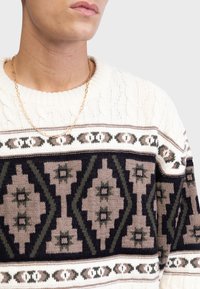 Mand iført en hvid kabelstrikket sweater med sorte og brune geometriske mønstre samt en tynd guldkæde.