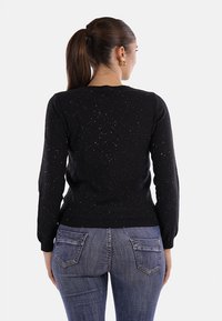 Donna con coda di cavallo castana che indossa un maglione a maniche lunghe nero a pois e jeans blu, in piedi di spalle contro uno sfondo bianco.
