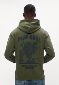 Olive zelená mikina s kapucňou s grafikou na chrbte zobrazujúcou kreslenú postavičku, textom "PLAY COOL" a rebrovanými manžetami a spodným lemom.