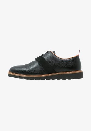 Chaussure oxford en cuir noir avec une finition texturée, des accents en daim et une semelle brune contrastante. Elle présente un bout rond et un design à lacets.