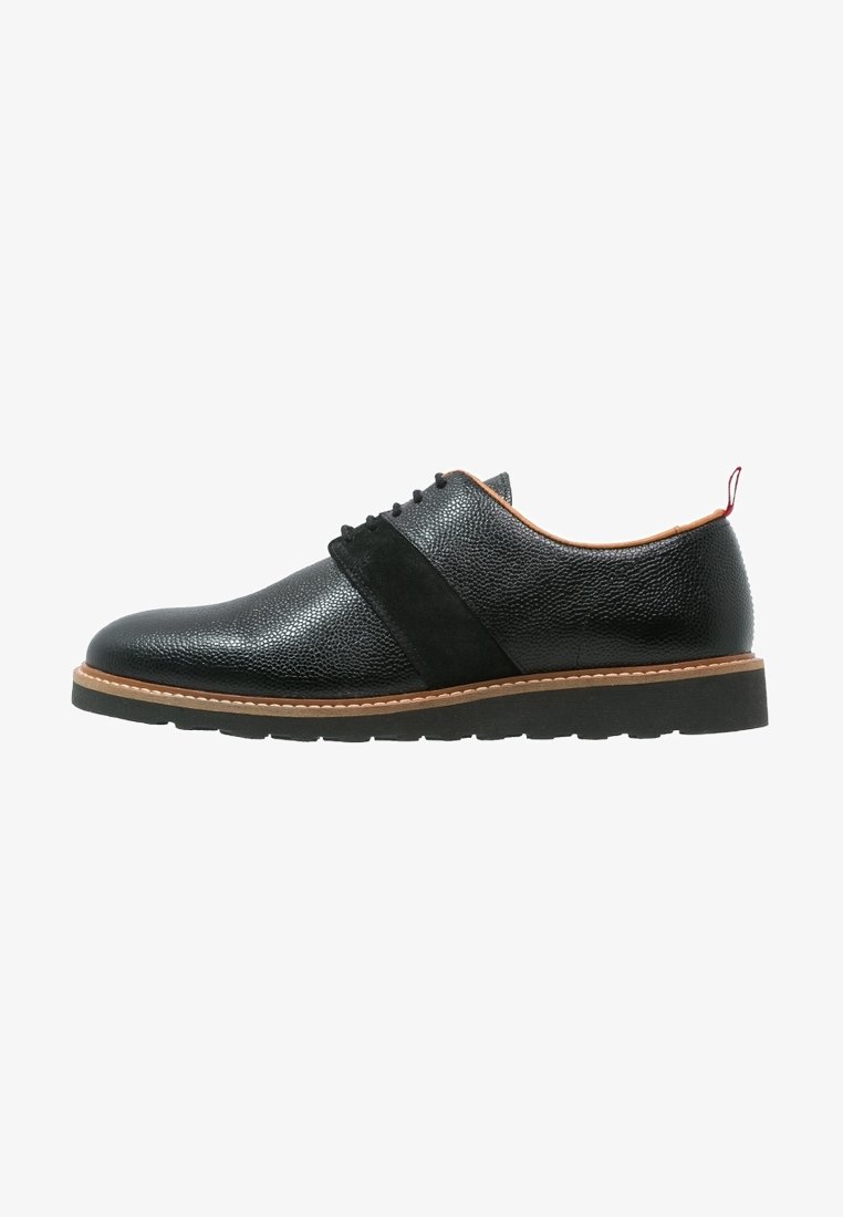 Chaussure oxford en cuir noir avec une finition texturée, des accents en daim et une semelle brune contrastante. Elle présente un bout rond et un design à lacets.