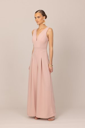 Žena v bezrukávovom, širokonohom overale ružovej farby s hlbokým výstrihom do V, doplnená kvetinovými náušnicami a zlatým náramkom.