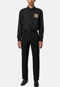Camicia nera a maniche lunghe con chiusura a bottoni e un emblema circolare dorato, abbinata a pantaloni neri coordinati e scarpe nere lucide.
