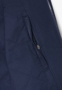 Tela acolchada azul marino con costuras diagonales que presenta una cremallera de bolsillo parcialmente abierta y una cremallera vertical adyacente en una chaqueta.