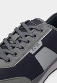 Κοντινή λήψη γκρι και ναυτικού μπλε αθλητικού παπουτσιού BOSS με υφασμάτινες και σουέντ υφές, γκρι κορδόνια και λογότυπο της μάρκας στην πλαϊνή επιφάνεια και στη γλώσσα.