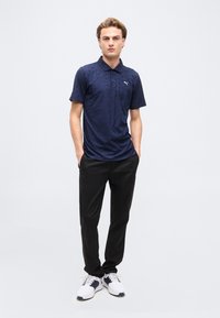 Jeune homme debout les mains dans les poches, vêtu d'un polo bleu marine, d'un pantalon noir et de baskets blanches, sur un fond uni.