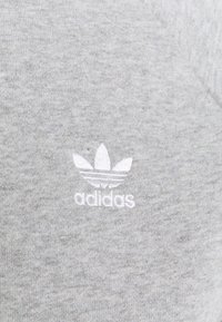 Grå bomullstyg med en vit broderad Adidas trefoil-logotyp med tre ränder. Mjuk textur och enhetlig färg genom hela tyget.