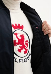 Chaqueta bomber de la marina parcialmente desabrochada sobre un suéter crema que presenta un emblema de león rojo y el texto azul "TOMMY HILFIGER". Detalle de punto texturizado visible.