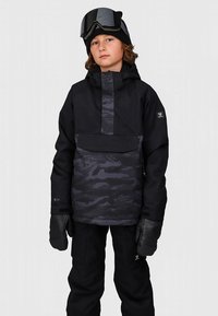 Fekete sídzseki terepmintás részletekkel, texturált ujjakkal, cipzáras zsebekkel, fekete kesztyűvel és símaszkkal párosítva, egy gyermek által viselve.