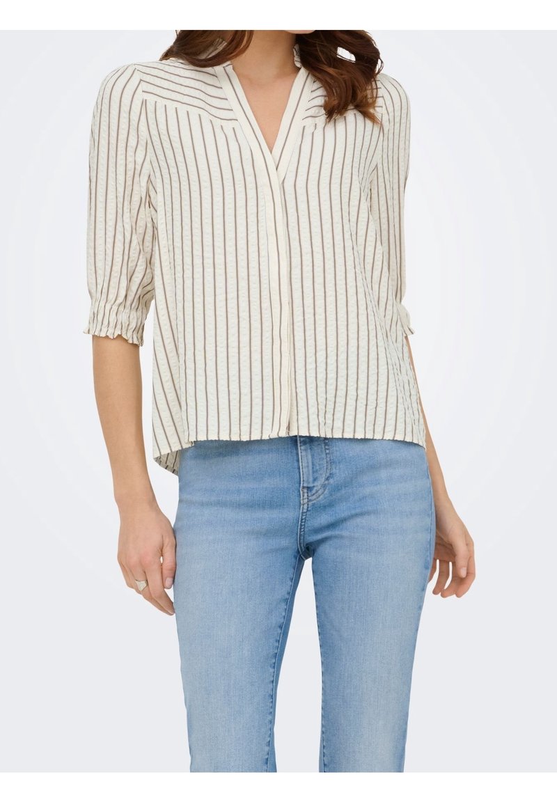 Femme portant un chemisier blanc à col en V avec des rayures verticales marron et des manches trois-quarts, associé à un jean bleu clair.