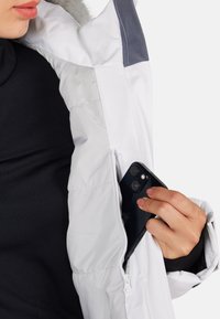 Osoba wkładająca czarny smartfon do wewnętrznej kieszeni zapinanej na zamek białej kurtki puchowej, nosząc czarną bluzkę pod spodem.
