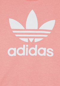 Tricou roz cu un mare logo Adidas alb, având trei dungi asemănătoare unor frunze deasupra numelui brandului, pe un fundal dintr-o țesătură texturată.