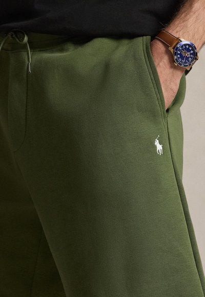 Vīrietis, kas valkā zaļas sporta bikses ar Polo logo, melnu augšdaļu un brūnu ādas pulksteni ar zilu ciparnīcu, roka kabatā.