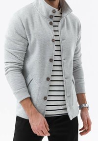 Chaqueta gris claro con cremallera y cuello alto, con botones marrones, bolsillos laterales y puños acanalados, superpuesta a una camiseta de rayas en blanco y negro.