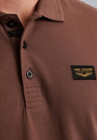 Hnedá polo tričko s textúrovanou tkaninou, s golierom, dvoma čiernymi gombíkmi a označením PME Legend so žltým textom a krídlovým emblémom.