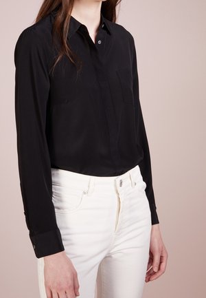 Femme portant une chemise noire à manches longues boutonnée, rentrée dans un jean blanc taille haute, debout devant un fond beige uni.