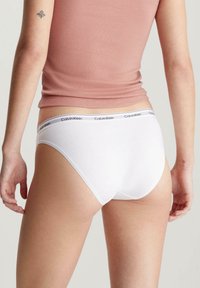 Slip in cotone bianco a vita bassa, con una cintura nera con logo. Design aderente con texture liscia e uno stile minimalista.