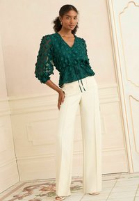 Blusa verde trasparente con trama floreale, scollatura a V e vita cintrata, abbinata a pantaloni bianchi a vita alta e gamba larga, in piedi su un pavimento decorato.