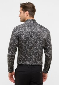 Man in een zwart bloemenlongsleeve shirt en zwarte broek, van achteraf gezien tegen een effen lichte achtergrond.