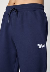 Tengerészkék jogger nadrág, puha anyagból készült, rugalmas derékkal és húzózsinórral, a bal combon egy fehér Reebok logóval.