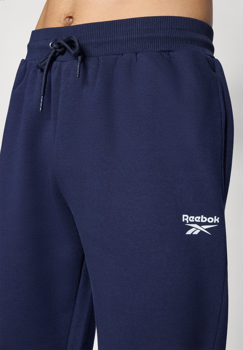 Pantalones jogging en color marino, confeccionados en un tejido suave, con cinturilla elástica y cordón de ajuste, y un logo de Reebok en blanco en el muslo izquierdo.