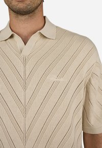 Beige kötött póló V nyakkal, átlós csíkokkal és nyitott szövésű mintákkal, rövid ujjakkal. Diszkrét logó részlet a mellkasán.