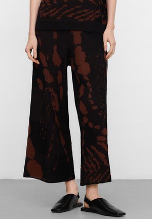 Schwarze Hosen mit weitem Bein und unregelmäßigen dunkelbraunen abstrakten Mustern, getragen mit schwarzen flachen Slingback-Schuhen.