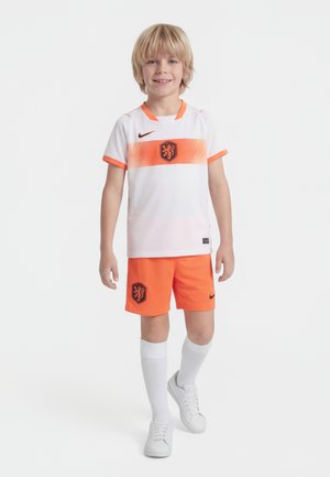 Jonge jongen lacht, draagt een wit en oranje voetbaluniform met leeuwemblemen, witte kniehoge sokken en witte sneakers, staand tegen een effen achtergrond.