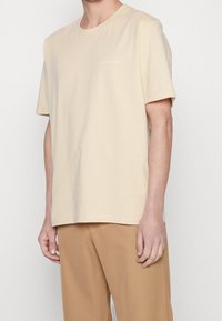 Man i en beige kortärmad t-shirt med texten "Tiger of Sweden" på bröstet och ljusbruna byxor mot en enkel bakgrund.
