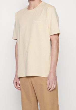Hombre con una camiseta de manga corta beige que lleva el texto "Tiger of Sweden" en el pecho y pantalones marrón claro, contra un fondo liso.