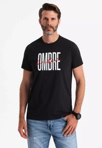 Camiseta de algodón negra con la palabra "OMBRE" en letras gruesas blancas y rojas, cuello redondo, mangas cortas, combinada con jeans rotos de color azul claro.