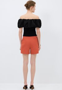 Top nero off-the-shoulder con maniche a sbuffo, abbinato a pantaloni corti arancioni su misura. La modella indossa scarpe con tacco nere e un bracciale dorato.