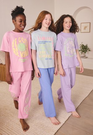 Trois enfants souriants marchant pieds nus à l’intérieur, portant des tenues décontractées aux couleurs pastel avec des messages positifs et des motifs floraux.