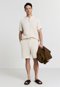 Uomo in camicia beige a maniche corte e pantaloni corti, che tiene in mano una giacca marrone, indossando sandali scuri intrecciati su uno sfondo chiaro e uniforme.