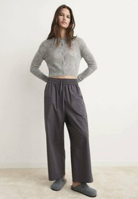 Vrouw die binnen staat in een grijze cropped cardigan, losse donkergrijze broek met touwtjes en grijze slippers op een tapijtvloer.