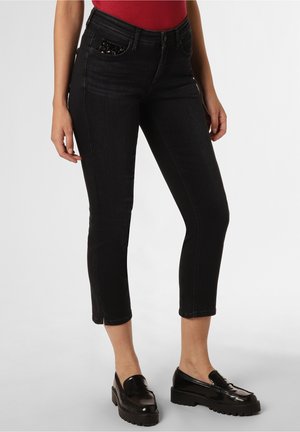 Vrouw draagt strakke zwarte cropped jeans met pailletten detail op de zak, zwarte plateau loafers en een rood topje, staand op een witte achtergrond.