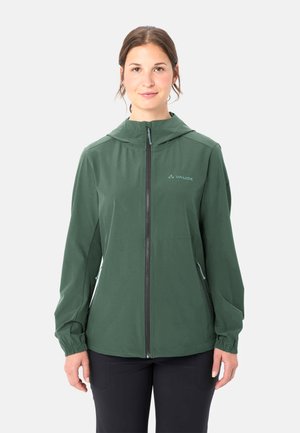 Frau steht und trägt eine grüne Kapuzenjacke mit Reißverschluss und schwarze Hosen vor einem schlichten hellen Hintergrund.