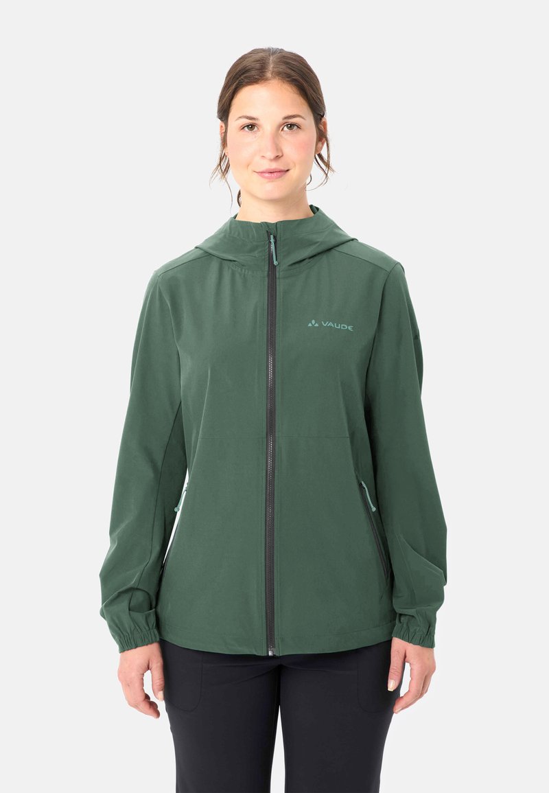 Donna in piedi che indossa una giacca verde con cappuccio e cerniera e pantaloni neri su uno sfondo chiaro e uniforme.