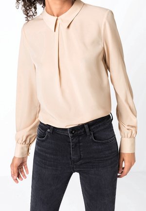Femme portant une blouse beige à manches longues avec devant plissé et col, associée à un jean noir taille haute sur fond blanc.