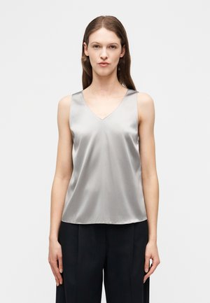 Sirgedpruunide juustega naine kannab hõbedast satiinist varrukateta V-kaelusega toppi ja musti laiade säärtega pükse, ta seisab ühevärvilise heleda tausta ees.