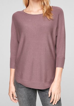 Femme portant un pull en maille mauve à manches trois-quarts et ourlet arrondi, assorti à un jean gris, sur un fond uni.
