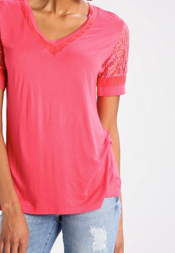Femme portant un t-shirt rose vif à col en V avec des manches courtes en dentelle et un jean bleu clair déchiré sur un fond uni.
