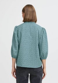 Tyrkysová blúzka s textúrovanou tkaninou, s nafúknutými rukávmi a rozptýleným čiernym bodkovaným vzorom. Obsahuje golier a nariadené manžety.