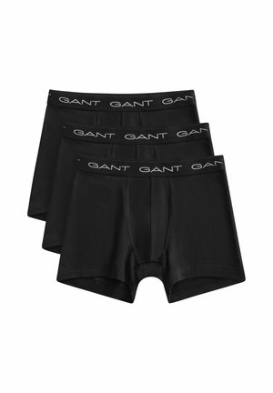 Drie boxershorts voor mannen in het zwart opgestapeld, elk met een zwarte tailleband waarop het witte "GANT"-logo herhaaldelijk is gedrukt.