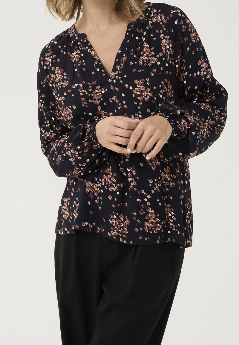 Femme portant un chemisier noir à manches longues avec un petit imprimé floral rose et blanc, associé à un pantalon noir ample, mains doucement jointes.