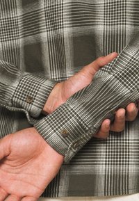 Grün-weiß kariertes Hemd mit knöpfbaren Manschetten und weicher Textur, zeigt sichtbare Karomuster und kontrastierende dunkle Bereiche.