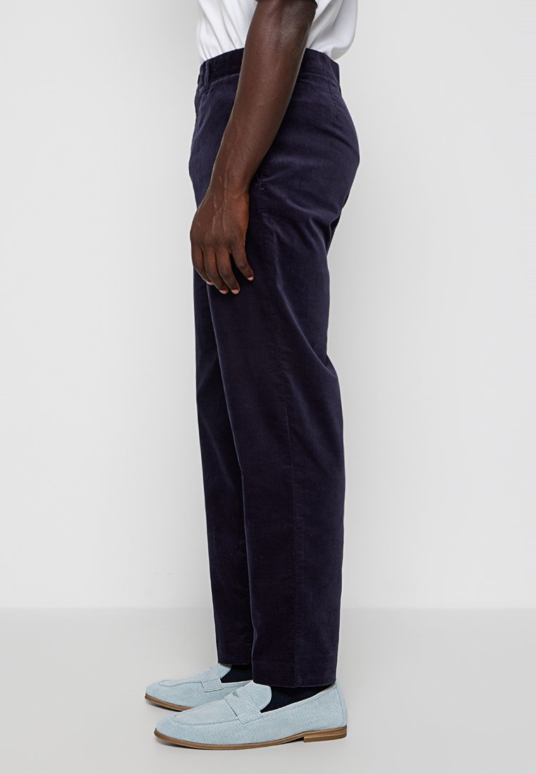 Man in donkere marineblauwe broek, een wit shirt, zwarte sokken en lichtblauwe instappers, staand tegen een effen achtergrond.