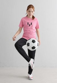 Noor, kellel on punased juuksed, kannab roosa särki ja musti pükse ning pallib must-valget jalgpalli siseruumides, tavalise seina taustal.